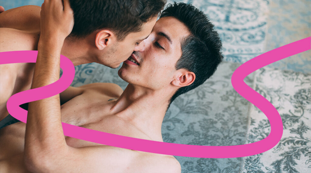 Two shirtless men kiss, embracing in a room with a patterned background and pink decorative ribbons.
