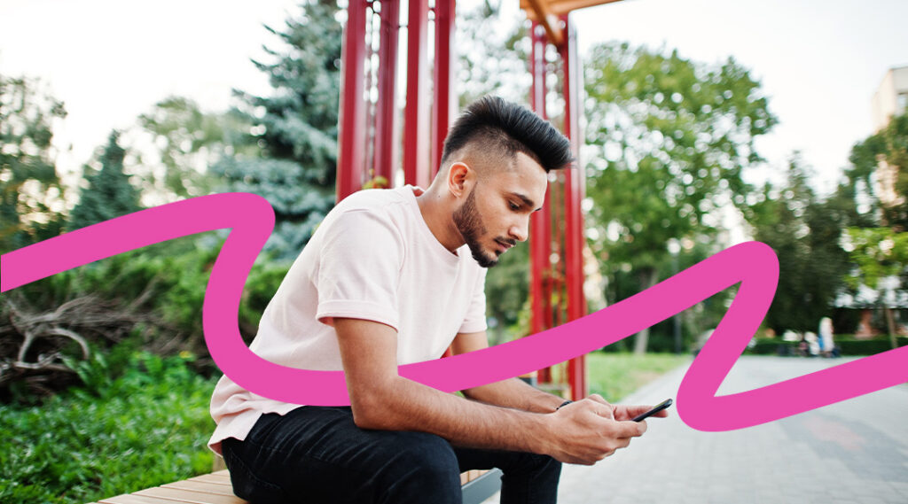 Man with a fade haircut sits on a bench in a park, focused on his smartphone as greenery and a red structure loom in the background, with a pink swoosh across the scene.